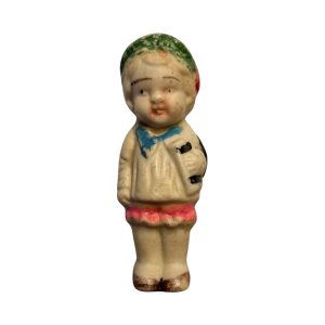Vintage Japanese bisque child figurine from the 1950s with cream outfit, pink trim, and green cap, showing age-related paint wear.